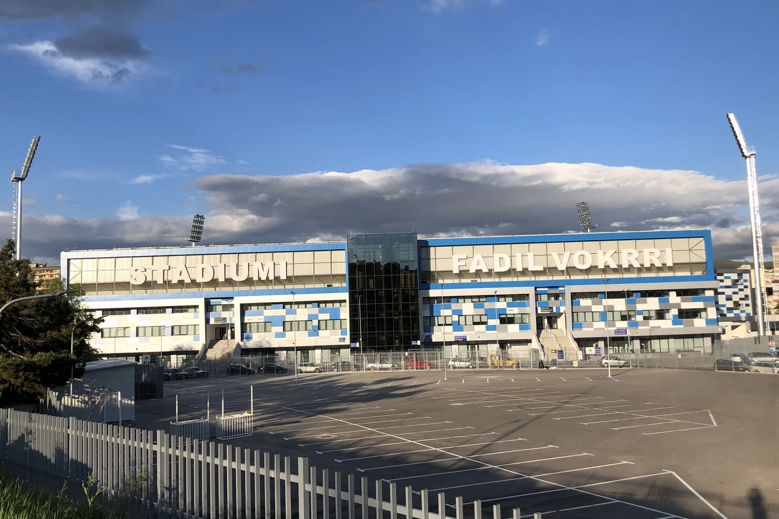 Γαλανόλευκη πρόσοψη του Stadiumi Fadil Vokrri με την ονομασία του σταδίου, προβολείς και χώρο στάθμευσης, σε περικομμένη φωτογραφία αρχείου.