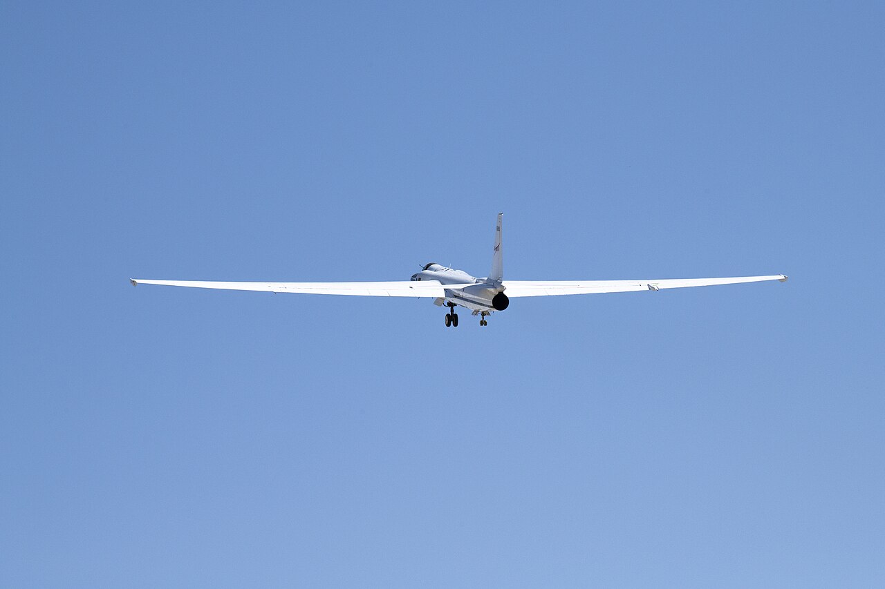 Λευκό αεροσκάφος ER-2 με ολόκληρο το άνοιγμα των πτερύγων και κατεβασμένο σύστημα προσγείωσης, από πίσω στον γαλάζιο ουρανό· αρχείο 2022.