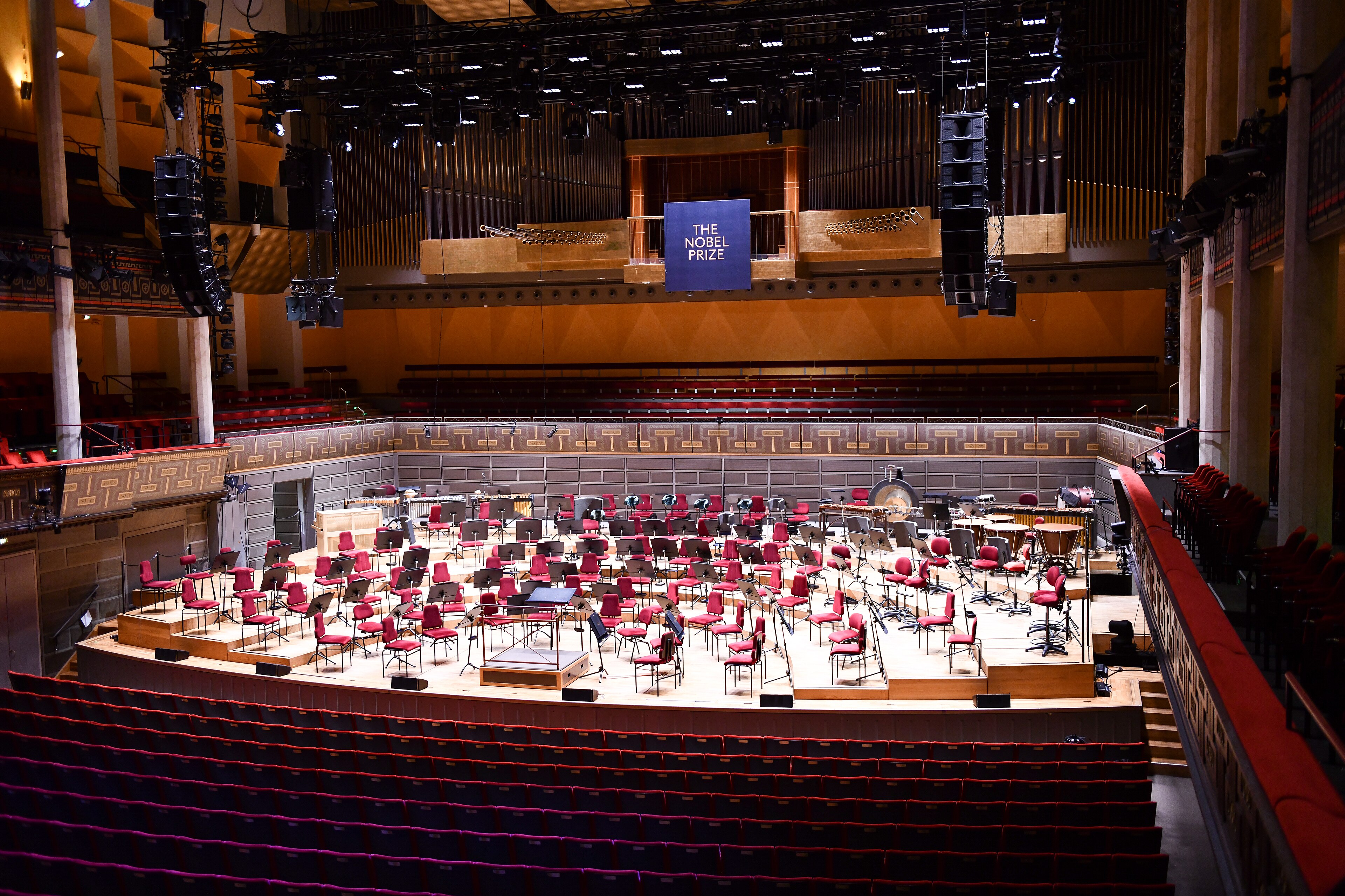 Άδεια σκηνή στο Konserthuset Stockholm με κόκκινες καρέκλες ορχήστρας, αναλόγια και επιγραφή Nobel Prize· αρχείο Δεκεμβρίου 2025.